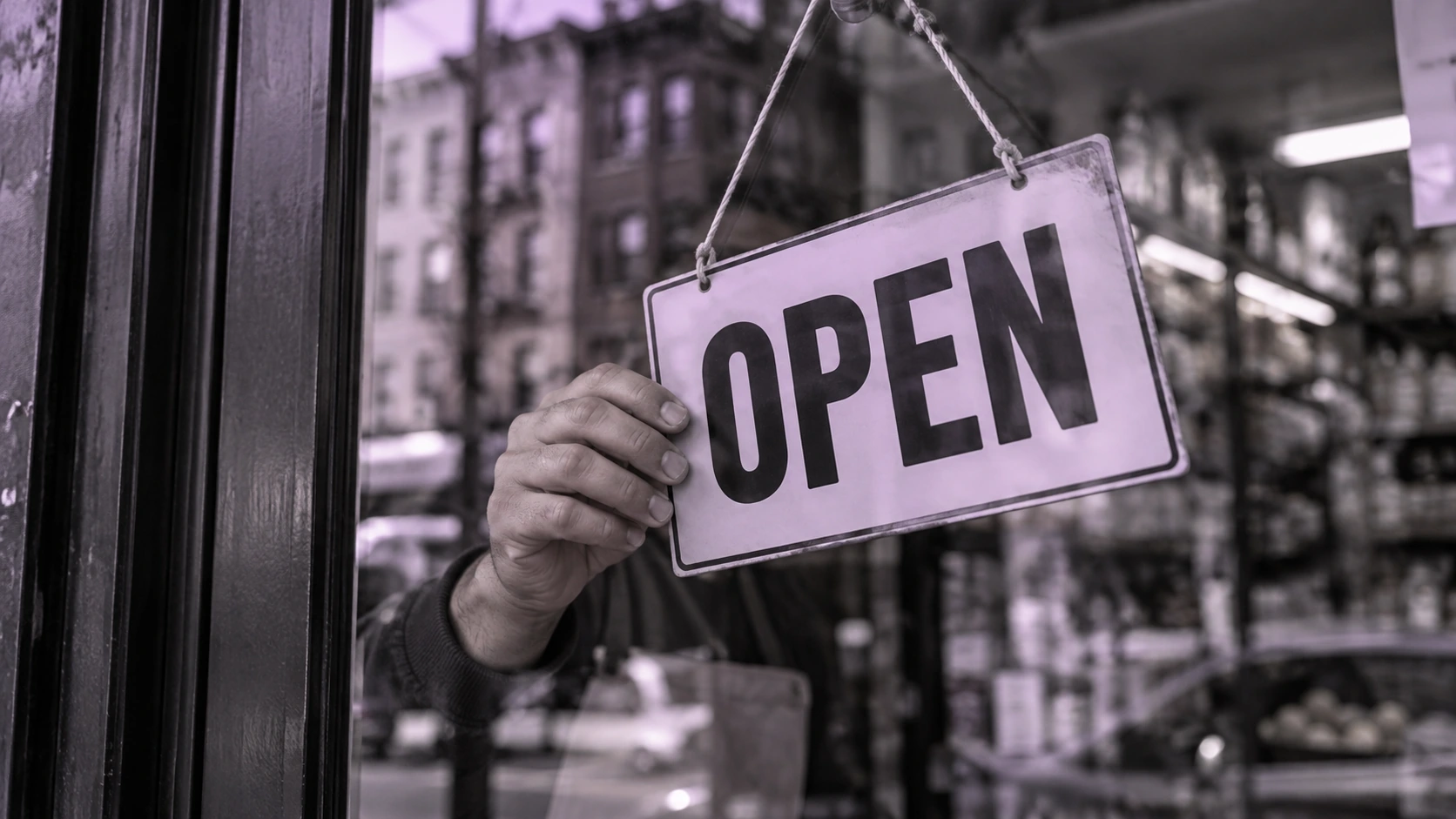 A hand holding an OPEN sign in a bodega door, with the dark door frame and street reflections visible in black and white with a restrained lavender tint
